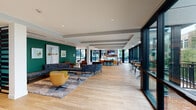This single-story clubhouse in Spring, TX is a thoughtfully designed community gathering space that blends contemporary aesthetics with functional versatility. The interior is anchored by light hardwood flooring that extends throughout, complemented by coffered ceilings with recessed lighting that lend architectural dimension to the open layout.

The main lounge area is a standout feature, defined by a bold teal accent wall adorned with curated artwork. Curved sectional sofas, leather armchairs, and vibrant accent ottomans create an inviting, social atmosphere. Floor-to-ceiling glass windows and sliding doors wrap the perimeter, flooding the space with natural light while offering unobstructed views of the surrounding courtyard and landscaped grounds.

A bar-height seating area with counter stools provides a casual alternative for gathering or working, while a designated lounge nook near the glass corner wall features a wall-mounted TV, a fireplace with a live-edge wood mantle, and comfortable seating. A private meeting or dining room offers a round wood table, modern chandelier, wall-mounted display, and direct access to the outdoor terrace.

A compact kitchenette serves the space with a stainless steel sink, wood cabinetry, and a built-in coffee station set against a striking geometric-patterned tile backsplash.