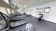 This fitness center offers a clean, functional environment designed to support a variety of cardiovascular workouts. The space is defined by its white-painted exposed beam ceiling, which adds architectural character while contributing to an open, airy feel. Ceiling fans are mounted overhead, promoting air circulation throughout the room.

The flooring consists of durable dark rubber gym matting, providing both safety and comfort underfoot. Along one wall, full-length mirrors visually expand the space and allow users to monitor their form during exercise. Natural light filters in through windows, brightening the interior alongside the room's existing lighting.

The facility is equipped with multiple treadmills arranged in a row, a recumbent exercise bike with an integrated display, a rowing machine, and additional cardio equipment. A wall-mounted air conditioning unit ensures climate control during workouts. Informational fitness posters and exercise guides are displayed along the walls, offering useful reference material for users. A large potted plant near the entrance adds a touch of greenery to the otherwise athletic setting, contributing to a welcoming atmosphere.