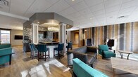Located at 6142 Merlot Dr in Highland, NY, this generously sized single-floor facility offers a thoughtfully designed layout spanning an impressive footprint. The welcoming hallways feature tiled flooring with inlaid detailing, coffered ceilings with recessed lighting, and comfortable seating areas that set an inviting tone throughout the space. The central lounge and bar area serves as a social hub, showcasing hardwood-style flooring, white decorative columns, a stone-accented bar with pendant lighting, and an eclectic mix of teal and charcoal upholstered seating. Adjacent to the lounge, a well-appointed dining area offers dark wood tables and upholstered chairs beneath large windows that frame lush outdoor greenery, filling the room with natural light. Multiple event and meeting rooms provide flexible gathering spaces, each equipped with built-in audio-visual capabilities, projection screens, and rows of seating suitable for presentations or group events. One room features a raised wooden platform stage and wall-mounted speakers, ideal for performances or lectures. A cozy library nook lined with dark wood bookshelves and comfortable reading chairs rounds out the amenities. Throughout the facility, a consistent palette of warm neutrals, modern fixtures, and refined architectural details creates a polished and functional communal environment.