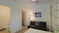 This single-floor space offers a clean, minimalist aesthetic defined by neutral white walls and light carpeted flooring that create a bright, welcoming atmosphere. The main living area is efficiently arranged, featuring a black tufted leather futon accented with decorative black-and-white patterned pillows, providing both seating and flexible sleeping options. A tall floor lamp with an adjustable reading light adds layered illumination to the space, complementing the overhead ceiling fixture with pendant-style drop lights that contribute a modern design element. A monochrome art print displayed on the wall adds a touch of personality and visual interest. The room connects directly to an adjoining space through a doorway, suggesting a functional flow between areas. Visible ceiling HVAC vents indicate a central air system throughout. The overall ambiance is understated and practical, with a neutral palette that offers a versatile backdrop suitable for various interior styles. The space is well-suited for those seeking a straightforward, low-maintenance living environment with modern conveniences.