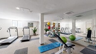 Located at 3205 W Floyd Ave in Denver, Colorado, this well-equipped fitness center offers a comprehensive workout facility across a single open floor. The space is designed with a clean, modern aesthetic, featuring neutral-toned carpet tiles in a grid pattern that define distinct workout zones throughout the room.

The cardio section includes multiple treadmills and an elliptical trainer, complemented by natural light from windows along the walls. A dedicated strength training area houses a multi-function cable machine, a rowing machine, a recumbent bike, and an adjustable weight bench, offering a full range of resistance-based exercises. A freestanding dumbbell rack provides a variety of free weight options for targeted training.

For functional fitness and flexibility, the space features a well-stocked medicine ball rack with an assortment of weighted balls, stability and exercise balls, and blue SmartMat exercise mats laid out for stretching or bodyweight workouts. Foam rollers and resistance accessories round out the functional training offerings.

Large floor-to-ceiling mirrors along one wall visually expand the space and allow users to monitor their form during workouts. Recessed ceiling lighting ensures the room is well-lit throughout. Potted plants are thoughtfully placed to add a touch of greenery and warmth to the otherwise crisp, white interior. An informational signage board is mounted near the entrance, completing this practical and inviting fitness environment.