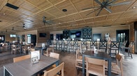 Located at 100 Foxborough Way in Richmond, Kentucky, this single-floor bar and dining venue offers a generously sized, open-concept interior designed with a warm yet upscale rustic aesthetic. The space is unified by a striking wood coffered ceiling fitted with multiple oversized ceiling fans, recessed lighting, and mounted flat-screen televisions throughout.

The main dining area features a mix of standard and high-top tables with cross-back bar stools and casual wooden chairs, all arranged to maximize capacity while maintaining a comfortable, social atmosphere. A full bar anchors one end of the space, finished with a stone face and flanked by dark cabinetry with X-pattern wine storage displays, creating a refined focal point.

At the opposite end, a relaxed lounge area offers leather and upholstered sofas, accent chairs, and coffee tables arranged over a vibrant area rug — ideal for more informal gatherings. A stone fireplace with a mounted TV above adds to the cozy ambiance of this zone.

Natural light floods the interior through floor-to-ceiling glass panel doors that line the rear wall, opening to views of an outdoor pool and surrounding community. Horizontal wood shiplap paneling and exposed brick accents add texture and warmth throughout, while polished concrete floors provide a durable, modern contrast to the rustic elements. The overall atmosphere is inviting, social, and well-suited for dining, entertaining, and community gatherings.
