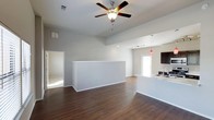 This two-story home offers a thoughtfully designed open-concept layout that promotes a sense of spaciousness and effortless flow between living areas. The main living space is anchored by striking dark hardwood flooring that extends throughout, complementing the clean, neutral tones of the walls and trim. Vaulted ceilings rise overhead, enhancing the airy, expansive feel of the room, while a ceiling fan with integrated lighting adds both comfort and practicality. A half-wall partition provides a subtle separation within the open floor plan, defining distinct zones without sacrificing the sense of openness. The kitchen is integrated seamlessly into the living area and features a light-colored countertop, a sink, and dark cabinetry accents that add a modern contrast. Pendant lighting in a bold red hue hangs above, serving as a distinctive design statement. Track lighting complements the kitchen space further, ensuring ample illumination. Natural light filters in through windows and a glass-paneled door, brightening the interior and creating a welcoming ambiance. Additional rooms are accessible from the main living area, and the two-story layout offers further living space above, making this a versatile and well-appointed home.
