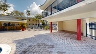 Located in Miami, FL along SW 65th Ave, this outdoor community pool area offers a well-appointed recreational retreat designed for relaxed Florida living. The centerpiece is a generously sized freeform swimming pool with a flowing, curved shape, featuring graduated depths clearly marked for safety. The pool's clear turquoise water is complemented by a wide, paver-tiled deck that provides ample space for lounge seating and casual outdoor gatherings.

A covered pavilion with a distinctive pagoda-style roof anchors one end of the pool area, offering shaded seating beneath its canopy. A vibrant mural decorates the pavilion wall, adding a colorful, artistic focal point to the space. Tall shade sails provide additional sun coverage over seating areas, enhancing comfort throughout the day.

The area is enclosed by a wrought iron security fence and is surrounded by mature tropical trees, including large oaks and palm trees, which provide natural shade and a lush, verdant backdrop. Adjacent low-rise residential buildings with warm yellow exteriors and covered balconies frame the space, contributing to an intimate and cohesive community atmosphere. The brick-paved courtyard areas extend the usable outdoor space, and covered parking beneath one of the buildings adds functional convenience to the overall property.