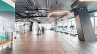 Located at 777 E Ocean Blvd in Long Beach, California, this contemporary fitness facility offers a well-appointed, expansive workout environment designed to accommodate a wide range of exercise needs. The main floor features durable tiled flooring throughout and is equipped with a full lineup of cardio machines—including treadmills and stair climbers—alongside a comprehensive selection of strength training equipment and cable machines. A dedicated studio space features mirrored walls, a ballet barre, and large floor-to-ceiling windows that flood the area with natural light and offer views of the surrounding palm tree-lined neighborhood. The industrial-style exposed ceiling is outfitted with striking hexagonal LED light fixtures, exposed ductwork, and conduit, lending the space a modern, urban aesthetic. Double glass entry doors provide a welcoming entrance, while wall-mounted displays add functional amenity. Adjacent to the fitness floor, an outdoor rooftop terrace extends the space with a circular lawn, communal dining seating with string lights, bold blue planters, and open views of the Long Beach skyline—creating an inviting setting for relaxation and outdoor gatherings.