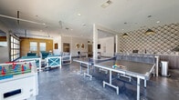 Located at 201 Telluride St in San Marcos, Texas, this single-floor community amenity space offers a generous, open-concept layout designed for recreation and socializing. The main living area features large-format dark tile flooring that flows seamlessly throughout, complemented by crisp white walls and recessed lighting that keep the space feeling bright and airy.

The expansive game and lounge area serves as the centerpiece of the space, accommodating a full-size Brunswick ping-pong table and a billiards table, both positioned beneath stylish pendant lighting. A dedicated lounge nook features comfortable seating, including gray sofas and white-framed teal accent chairs, arranged around a wall-mounted flat-screen TV and a warm mustard-yellow accent wall with natural light filtering in through multiple windows. An eye-catching geometric patterned wall adds a bold design element near the game area.

Large industrial-style glass garage doors open the space to the outdoors, flooding the interior with natural light. A wet bar with a white countertop and subway tile backsplash provides a functional serving area for gatherings.

Two restrooms are finished with classic white subway tile walls, dark stone-look flooring, private stalls, and wall-mounted vanities with mirrors, offering a clean and functional design consistent with the overall modern aesthetic of the space.