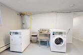 Located at 9 Amos Ave in Waterloo, Ontario, this single-floor utility space provides a range of practical amenities across a compact but well-organized footprint. The layout includes an entrance corridor, a shared laundry area, dedicated storage lockers, a boiler room, and an electrical room.

The entrance corridor is brightly lit with overhead fluorescent lighting and features durable grey flooring throughout, lending the space a clean, functional aesthetic. White-painted walls keep the interior bright and easy to maintain. Signage clearly marks the boiler and electrical rooms, which are accessible via the main hallway.

The laundry area is equipped with commercial-grade Huebsch washers and dryers, a stainless steel utility sink, and a folding/sorting table, making it well-suited for shared residential use. A wall-mounted bulletin board provides a space for notices and instructions. Gas supply lines and ventilation ductwork are visible overhead, reflecting the utilitarian nature of the space. A window in the laundry room allows for natural light.

A separate storage area features a row of numbered individual storage lockers with slatted ventilation panels above, offering secure personal storage for multiple users. The grey epoxy-style flooring is consistent throughout, contributing to a clean and practical environment overall.