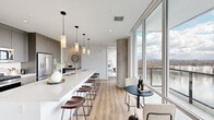 This well-appointed 2-bedroom, 3-bathroom residence showcases a contemporary design defined by floor-to-ceiling windows that frame sweeping panoramic views of the surrounding cityscape and waterway. Light wood flooring flows throughout the single-level layout, complementing the clean, modern aesthetic.

The open-concept kitchen and living area serves as the heart of the home. The kitchen features an elongated white quartz island with bar seating, gray shaker cabinetry reaching toward the ceiling, stainless steel appliances, and pendant lighting. The adjacent living area benefits from an exposed concrete ceiling and structural columns, adding an industrial architectural contrast to the otherwise bright, white interiors. Floor-to-ceiling glazing floods the space with natural light and provides direct access to a balcony.

Both bedrooms are generously proportioned, with the primary bedroom offering expansive window views of the water and skyline. Each bedroom connects to its own bathroom, with the primary bath featuring a double-sink vanity with quartz countertops, gray cabinetry, and a soaking tub. Two dedicated walk-in closets provide substantial storage with built-in shelving and hanging rods throughout.