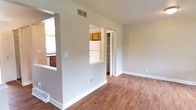 This three-floor residence offers a practical and well-organized layout with distinct living zones connected by thoughtful architectural details. The main living area features hardwood-style flooring and neutral gray walls, creating a clean, versatile backdrop. A distinctive pass-through window cutout in the dividing wall visually connects the living space to the kitchen, enhancing the sense of openness while maintaining defined areas. A carpeted staircase is conveniently accessible from the main level, providing access to the upper floors.

The kitchen is equipped with warm honey-toned wood cabinetry both above and below the counters, complemented by dark granite-style countertops. A double stainless steel sink sits beneath a large window that frames views of mature trees and the surrounding neighborhood, allowing ample natural light into the space. White appliances, including a range and dishwasher, complete the functional kitchen setup.

Adjacent to the living area, a closet with bifold doors provides convenient storage. The overall design maintains a consistent neutral palette throughout, with white trim and baseboards accenting each room. The multi-floor layout offers well-separated living, sleeping, and utility spaces suited for everyday comfort.