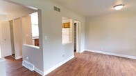This three-floor residence offers a practical and well-organized layout with distinct living zones connected by thoughtful architectural details. The main living area features hardwood-style flooring and neutral gray walls, creating a clean, versatile backdrop. A distinctive pass-through window cutout in the dividing wall visually connects the living space to the kitchen, enhancing the sense of openness while maintaining defined areas. A carpeted staircase is conveniently accessible from the main level, providing access to the upper floors.

The kitchen is equipped with warm honey-toned wood cabinetry both above and below the counters, complemented by dark granite-style countertops. A double stainless steel sink sits beneath a large window that frames views of mature trees and the surrounding neighborhood, allowing ample natural light into the space. White appliances, including a range and dishwasher, complete the functional kitchen setup.

Adjacent to the living area, a closet with bifold doors provides convenient storage. The overall design maintains a consistent neutral palette throughout, with white trim and baseboards accenting each room. The multi-floor layout offers well-separated living, sleeping, and utility spaces suited for everyday comfort.