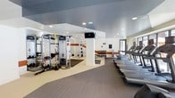 This contemporary fitness center offers a comprehensive workout environment designed to accommodate a wide range of exercise routines. The space is thoughtfully organized into distinct functional zones, allowing users to move seamlessly between cardio, strength, and functional training areas. Along one side of the room, a row of treadmills provides ample cardio capacity, positioned to make the most of the available floor space. A well-stocked dumbbell rack sits centrally, offering a full range of free weights alongside a dedicated flat bench for strength training. The functional training zone features a multi-station cable and resistance system, ideal for full-body workouts, complemented by TRX suspension straps and a squat or power rack for more advanced training. A wall-mounted flat-screen television adds a modern amenity for entertainment during workouts. The flooring transitions between rubberized matting in the training zones and durable gym-grade carpet, ensuring both safety and comfort throughout. Recessed lighting in the smooth ceiling keeps the entire space bright and welcoming, while large windows allow natural light to filter in. Clean white walls accented with warm orange striping give the gym a polished, energetic aesthetic. Overall, the fitness center presents a professional and inviting atmosphere suitable for all fitness levels.