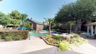 This single-story community amenity complex is defined by its inviting resort-style outdoor environment and classic brick construction. At the heart of the property lies a spacious swimming pool with a separate spa, both framed by natural stone retaining walls and lush, meticulously maintained landscaping. Tall palm trees, mature shade trees, colorful flowering plants, and manicured hedgerows create a verdant, tropical atmosphere throughout the grounds.

The pool deck is generously sized, providing ample room for lounge chairs and shaded seating areas, ideal for relaxing under open skies. A connected spa area sits at an elevated tier, accessed by stone steps and bordered by layered stonework planters filled with vibrant seasonal blooms.

The clubhouse building features traditional red brick exterior walls, large multi-pane windows that allow abundant natural light, and a covered walkway with white columns running along its perimeter. This covered corridor leads to the on-site fitness center, clearly marked and accessible directly from the outdoor patio. Additional outdoor seating is thoughtfully arranged near the building's entrance. Surrounding multi-story residential buildings with brick and siding exteriors complete the well-established community setting.