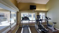 This single-floor fitness center is designed to provide a functional and comfortable workout environment. The room features a vaulted ceiling with angled beams that add architectural interest while enhancing the sense of vertical space. Warm golden-yellow walls create an energizing atmosphere throughout the space.

Large windows line multiple walls, flooding the room with natural light and offering clear views of an outdoor pool and surrounding grounds. This connection to the outdoors adds an open, airy quality to the workout area.

The space is equipped with cardio machines, including a treadmill and an elliptical trainer, positioned to take full advantage of the pool-facing windows. A wall-mounted flat-screen television is prominently installed near the ceiling, allowing users to stay entertained during their workouts. A clock is also mounted nearby for convenience.

The flooring features a low-pile carpet suitable for exercise use, and a tiled ledge runs beneath the windows, adding a practical surface detail. Mirrored panels on one wall visually expand the space and allow users to monitor their form. Overall, the fitness center offers a well-lit, efficiently organized environment with thoughtful design elements that make for a pleasant and motivating exercise experience.