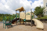 This outdoor playground area offers a dedicated recreational space designed for active play. The centerpiece of the space is a multi-level play structure constructed with durable materials, featuring tan slatted panel walls and green metal framing. A wooden-rung climbing ladder provides access to the elevated platform, encouraging physical activity and play. The structure is supported by sturdy brown metal posts and is set atop a gravel surface, which serves as a safety-conscious ground cover across the entire play area. The playground is enclosed by a black metal fence, clearly defining the boundaries of the space and providing a secure environment. Raised stone retaining walls border one edge of the area, adding a structured landscaping element along with green shrubbery that softens the surroundings. The space is situated adjacent to a multi-story building with a light-colored exterior, whose windows overlook the play area. The open-air setting allows for ample natural light and an unobstructed view of the sky, creating a bright and welcoming atmosphere. Overall, this playground provides a functional and well-maintained outdoor amenity suited for recreational use.