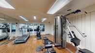 Located at 23825 Anza Avenue in Torrance, California, this fitness center offers a thoughtfully designed single-floor workout space that is both functional and inviting. The room features warm hardwood flooring throughout, white vertical shiplap-style wall paneling, and a full-length wall mirror that visually expands the space and supports proper form during exercise. Overhead, recessed skylight panels and fluorescent lighting provide consistent, bright illumination across the entire area. The cardio section is well-appointed with multiple treadmills, an elliptical trainer, and upright stationary bikes, positioned to take advantage of natural light from large windows overlooking an adjacent outdoor area. The strength training section includes a multi-function cable machine, adjustable weight benches, free weights, and a wall-mounted shelf for accessories. Stability balls and medicine balls round out the functional fitness offerings. The clean, neutral color palette and open floor plan create an uncluttered, motivating atmosphere suitable for a variety of workout routines. Large glass panels along one wall allow views to the exterior, adding a sense of openness. This fitness center delivers a comprehensive and well-maintained environment for residents or users seeking convenient, on-site exercise facilities.