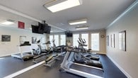 Located at 1047 Hwy 31 in Spanish Fort, Alabama, this single-floor fitness center is a well-equipped space designed to support a comprehensive workout experience. The room is lined with floor-to-ceiling mirrors along one wall, creating a sense of expanded space and allowing users to monitor their form during exercise. Large windows with plantation-style blinds line the opposite wall, flooding the interior with abundant natural light and offering views of the surrounding greenery. The facility is outfitted with a variety of cardio machines, including treadmills and elliptical trainers, arranged in an orderly fashion across a durable dark rubberized floor. A dedicated free weights area features a full dumbbell rack, along with a workout bench for strength training exercises. Multiple flat-screen televisions are mounted throughout the room — both on the mirrored wall and suspended from the ceiling — providing entertainment during workouts. Recessed and flush-mounted overhead lighting ensures the space remains bright and well-lit at all hours. Crown molding along the ceiling adds a refined architectural touch, elevating the overall aesthetic of the room. The layout is thoughtfully organized to maximize usable floor space while maintaining comfort and functionality for all fitness levels.