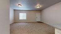 This two-story residence presents a clean, functional design throughout, finished in warm neutral tones that create a cohesive and inviting atmosphere. The main living area is generously proportioned, featuring plush wall-to-wall carpeting, textured ceilings, and dual flush-mount light fixtures that provide consistent illumination. A window with blinds allows natural light to fill the space, while a solid entry door with a modern lock adds a sense of security and structure.

The kitchen is efficiently designed with white shaker-style cabinetry, granite-look countertops, and black appliances including an electric range and over-the-range microwave. Wood-look flooring grounds the space and offers a practical contrast to the carpeted areas elsewhere.

The bathroom is well-appointed, featuring a white vanity with granite countertops, dark hardware accents, a single undermount sink, and a full tub-shower combination with built-in shelving. Tile flooring adds durability and a clean aesthetic.

A dedicated laundry room houses a full-size washer and dryer, tile flooring, overhead shelving, and an electrical panel for added convenience. Additional carpeted rooms offer versatile living or sleeping spaces, each with natural light from blinds-covered windows and consistent neutral wall finishes throughout.