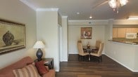 This single-floor residence offers a thoughtfully designed interior that balances comfort and elegance. The open layout creates a natural flow between the living and dining areas, making the space feel welcoming and functional. Warm, neutral wall tones provide a calm, inviting ambiance throughout, complemented by crisp white crown molding that adds a refined architectural detail to each room.

The dining area is a standout feature, centered around a round dark-wood table with cushioned beige chairs, offering an intimate setting for meals. A framed still-life artwork adorns the wall above, adding a subtle decorative touch. Recessed lighting overhead provides even, practical illumination throughout the space.

Rich, dark hardwood flooring runs throughout, grounding the interior with warmth and visual continuity. The living area transitions smoothly from the dining space, furnished with a comfortable sofa accented by warm-toned throw pillows, alongside a classic table lamp that contributes to the room's cozy atmosphere.

Architectural elements such as the defined column-style wall divisions add structure and character to the open floor plan without interrupting the sense of spaciousness. Ceiling vents ensure efficient climate control, while the clean, uncluttered design makes this a practical and pleasant space to inhabit.