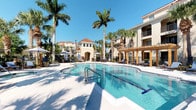 This inviting outdoor amenity space showcases a resort-style swimming pool as its centerpiece, finished with pristine white tile decking and shimmering blue water. The pool area is generously sized, offering dedicated lap lanes alongside a broader recreational swimming area, with easy access provided by stainless steel entry rails. Surrounding the pool, a spacious sun deck is furnished with lounge chairs and umbrellas, perfect for relaxation under the Florida sun. A handsome wooden pergola anchors one side of the space, providing a shaded retreat and adding a warm architectural contrast to the surroundings. A charming arched gatehouse or pavilion serves as a focal point in the background, framed by towering royal palms and lush tropical landscaping that enhance the resort atmosphere. The perimeter is enclosed by decorative black iron fencing, ensuring privacy and security. The Mediterranean-inspired residential buildings rise in the background, featuring terracotta tile rooflines, stucco exteriors, and private balconies that overlook the pool area. Abundant natural light and open blue skies complete the ambiance, making this outdoor space a vibrant and welcoming community amenity ideal for both active recreation and leisurely enjoyment.