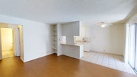 This single-floor space offers a clean, functional layout designed to maximize everyday livability. The living area features warm hardwood-style flooring that creates a welcoming atmosphere, while a textured popcorn ceiling adds a classic architectural touch. A set of built-in open shelves is integrated into the wall, providing convenient display or storage space without occupying additional floor area.

The kitchen is positioned adjacent to the living area and is defined by light ceramic tile flooring, offering a clear visual transition between spaces. A pass-through counter connects the kitchen to the living area, promoting an open and communicative flow. The kitchen is equipped with white cabinetry, a sink, and countertops that reflect the space's clean, neutral aesthetic. A ceiling fan in the kitchen area aids in ventilation and comfort.

The overall color palette is bright and neutral, with white walls and ceilings that help the space feel open and airy. The layout is straightforward and well-organized, making efficient use of every area. Natural light contributes to the inviting atmosphere, while the minimal design allows for flexible furnishing and personalization throughout the home.