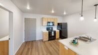 Located at 5340 W Vision Drive in Sioux Falls, South Dakota, Unit 205 is a thoughtfully designed single-floor residence that offers a clean, modern aesthetic throughout. The kitchen is a standout space, featuring warm honey-toned cabinetry paired with white countertops, durable wood-look laminate flooring, and black appliances including a refrigerator, range with microwave above, and a double-basin sink. Recessed lighting and a pendant light over the counter provide ample illumination, while an open layout creates an easy flow for everyday living. The bedroom is bright and comfortable, finished with plush carpeting and a window that allows natural light to filter in, offering views of the surrounding landscape. The full bathroom is efficiently laid out and divided into distinct functional zones — a dual-sink vanity with warm wood cabinetry and a large mirror is complemented by a separate water closet housing the toilet, and a walk-in shower. Tile-look flooring runs throughout the bathroom, adding a clean and cohesive finish. The overall design of the unit is straightforward and practical, with neutral tones, white walls, and consistent finishes that provide a versatile backdrop for any furnishing style.