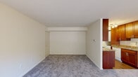 This efficient one-bedroom, one-bathroom unit is thoughtfully laid out across a single floor, making the most of its cozy footprint. The living room is the central gathering space, featuring soft wall-to-wall carpeting, neutral cream-toned walls, and a front entry door flanked by two windows that invite natural light into the room. A structural beam detail adds subtle architectural character to the ceiling. The galley-style kitchen is well-appointed with rich cherry wood cabinetry, granite-patterned countertops, a stainless steel sink, and white appliances including a gas range, over-the-range microwave, and refrigerator. Hardwood-style flooring runs through the kitchen, complementing the warm cabinetry tones. The bedroom offers a comfortable and private retreat, carpeted underfoot, with a window providing natural light and a closet for storage. The bathroom is clean and functional, featuring a single-sink vanity with a wood cabinet base, a full tub-shower combination with white tile surround, and classic tile flooring. Throughout the unit, the consistent use of neutral tones and warm wood accents creates a cohesive and livable atmosphere.