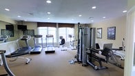 This fitness room is designed to support a well-rounded workout routine, offering a thoughtful selection of both cardio and strength training equipment. The space features multiple treadmills and an upright stationary bike positioned along a row of large, arched windows dressed with dark valances, allowing generous natural light to fill the room while providing views to the outdoors. A multi-station cable and weight machine anchors the strength training area, complemented by a leg press machine, making the space suitable for a full-body workout. The room is finished with neutral-toned carpeted flooring and smooth walls in a warm beige palette. Recessed lighting across the ceiling ensures consistent, even illumination throughout. Motivational fitness posters and exercise reference charts are displayed on the walls, adding a functional and informative touch to the décor. The layout is open and well-organized, allowing users to move comfortably between equipment. Overall, the fitness room presents a clean, practical, and welcoming environment suited for daily exercise.