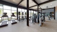 This fitness center delivers a polished, professional workout environment designed to accommodate a wide range of exercise needs. The space is defined by its striking floor-to-ceiling windows along the perimeter, flooding the interior with natural light and offering open views of the surrounding outdoor landscape. Dark-framed structural columns punctuate the open floor plan, adding an industrial-modern architectural character that is complemented by neutral-toned flooring and recessed lighting throughout.

The main workout area is generously sized, housing an extensive array of equipment. Cardio offerings include treadmills, ellipticals, rowing machines — including a Concept2 rower — and stationary bikes. A dedicated strength training section features Matrix cable machines, cable crossover systems, weight benches, and a full dumbbell rack with a broad range of free weights. Large wall mirrors amplify the sense of space and allow for form monitoring during workouts.

The entry area features warm wood-panel wall accents, glass doors, a water fountain station, and built-in storage cubbies, creating a welcoming transition into the facility. Ceiling fans and recessed lighting ensure comfort throughout. The overall ambiance is modern, clean, and motivating — a thoughtfully designed space built to support a complete fitness routine.