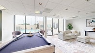 Located at 2828 Southwest Fwy in Houston, TX, this well-appointed community lounge offers a thoughtfully designed recreational space that balances social and leisure activities. The room is finished with light hardwood-style flooring, crisp white walls, and a drop tile ceiling with recessed and panel lighting that creates a bright, welcoming atmosphere.

A centerpiece pool table with a navy felt surface anchors the main area, making it ideal for casual gatherings. Adjacent to it, a comfortable lounge zone features a plush neutral-toned sofa, an ottoman, and accent chairs arranged over a vintage-style area rug, accompanied by a large wall-mounted flat-screen TV and a sleek media console. Curated artwork, including a dynamic equestrian painting and abstract circular canvases, adds visual interest throughout the space.

A dedicated café nook showcases a sage green accent wall adorned with two large circular art pieces, paired with round bistro tables and warm cognac leather-upholstered bar stools — perfect for casual conversation or working. Floor-to-ceiling glass walls and sliding doors on one side flood the room with natural light and provide direct views of and access to the outdoor pool and deck area, seamlessly connecting indoor and outdoor living. A potted tree adds a refreshing natural touch to the overall modern aesthetic.