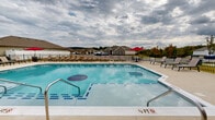 Located at 8919 Apison Pike in Collegedale, Tennessee, this outdoor community amenity area serves as a central hub for residents of Apison Village. The centerpiece of the space is a generously sized swimming pool with clearly marked depth gradations, ranging from a shallow entry zone to a deeper swimming area, making it suitable for a wide range of users. The pool features clean white coping and a branded pool medallion on the floor, reinforcing the community identity.

Surrounding the pool is a wide, open concrete sundeck outfitted with rows of lounge chairs, providing ample space for relaxation and sunbathing. Vibrant red patio umbrellas are positioned throughout the deck, offering shaded seating options for those seeking relief from the sun. Additional table-and-chair sets are arranged near the umbrellas, creating inviting social gathering spots.

Adjacent to the pool, a covered structure houses what appears to be restroom or changing facilities, accessed via bold black doors. An outdoor television is mounted under the covered overhang, enhancing the entertainment value of the space. The entire pool area is enclosed by a black wrought-iron style fence, ensuring safety and defined boundaries. Beyond the fence, well-manicured green lawns and single-story residential homes with neutral exteriors and pitched rooflines frame the backdrop, with rolling Tennessee treelines visible in the distance.