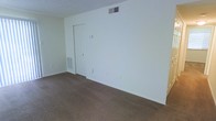 This single-floor space presents a straightforward, functional layout with a clean and neutral aesthetic throughout. The room is finished with wall-to-wall carpeting in a warm beige-brown tone, providing a soft underfoot feel that extends across the open floor area. The walls are painted in a crisp, bright white, creating a light and airy atmosphere that makes the space feel open and easy to personalize. Ceiling-mounted HVAC venting ensures climate control is integrated seamlessly into the design, while a matching floor-level vent further supports efficient air distribution throughout the room. A classic white paneled door provides access to an adjoining space or closet, featuring a simple and timeless design consistent with the overall neutral palette. The textured ceiling adds a subtle dimension to the overhead finish. Multiple electrical outlets are conveniently positioned along the walls, offering practical utility for furnishing and everyday use. The space is clean, move-in ready, and well-suited for a variety of uses, offering a blank canvas that allows for flexible interior design and arrangement.