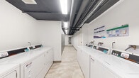 This shared utility space is designed to efficiently serve building residents, combining a fully equipped laundry room with a dedicated storage corridor on a single floor. The laundry area is lined with multiple commercial-grade front-loading washers and dryers, each equipped with digital payment interfaces for convenient operation. A built-in folding counter provides a practical workspace alongside the machines, and a wall-mounted mini-split unit ensures comfortable temperature control throughout the room. Informational signage is posted for user guidance.

The space is finished with light-toned tile flooring throughout, lending a clean and cohesive aesthetic. Exposed piping and ductwork run along the dark-painted ceilings, giving the space an industrial character while bright fluorescent strip lighting ensures excellent visibility. White walls contribute to an open, well-maintained feel.

Adjacent to the laundry room, a wide corridor is lined on one side with floor-to-ceiling individual storage lockers constructed from metal mesh, offering residents secure and ventilated personal storage. The layout flows logically from the laundry area through the storage corridor, terminating at a classic white-framed glass-pane door. A fire extinguisher is prominently mounted for safety. The overall space balances functional utility with a tidy, modern presentation.