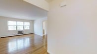 This single-floor space offers a clean, open layout defined by its bright, minimalist aesthetic. Polished hardwood floors run throughout, reflecting natural light and adding warmth to the interior. The crisp white walls create a fresh, airy atmosphere that enhances the sense of openness across the main living area.

The main living space is generously proportioned, with a wide, unobstructed floor plan that provides excellent flexibility for furnishing and use. A large window on the far wall invites an abundance of natural light, framing views of mature trees outside and contributing to the space's welcoming ambiance. A baseboard heating unit runs along the exterior wall, ensuring comfort throughout cooler months.

A structural soffit detail adds architectural interest to the transition between spaces, while a wall-mounted unit — likely a mini-split climate system — provides additional climate control options. An open doorway leads to an adjacent room, suggesting a well-connected and functional floor plan. The overall design is understated and neutral, making it highly adaptable to a variety of uses. The combination of natural light, hardwood flooring, and clean lines gives this space a timeless and practical character.