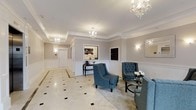 This single-floor space presents a beautifully appointed lobby area defined by its classic, upscale design aesthetic. The room is finished with warm greige walls accented by crisp white crown moulding and wainscoting, lending a timeless, formal elegance throughout. Underfoot, a polished marble-style tile floor with dark diamond inlays adds a refined architectural detail that anchors the space visually.

Overhead, multiple crystal chandeliers and recessed lighting illuminate the room with a warm, inviting glow, highlighting the generous ceiling height and the carefully curated interior. Wall sconces provide additional ambient lighting, enhancing the overall sense of warmth and sophistication.

The seating area features tufted blue upholstered armchairs arranged around a small side table, offering a comfortable and stylish gathering spot. A sleek console table is positioned along one wall beneath a framed artwork, while a built-in display or mailbox unit is recessed into the adjacent wall, suggesting the functional purpose of the space as a communal lobby or reception area.

A wide, well-lit corridor extends toward what appears to be a private entrance, reinforcing the sense of depth and spaciousness within the space.