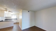 This single-floor residential space offers a clean, functional layout designed for comfortable everyday living. The interior is characterized by a neutral white and cream color palette across the walls, creating a bright and airy atmosphere. Dark hardwood-style flooring runs throughout the main living area, providing a warm contrast to the light-toned walls and adding a sense of depth to the space.

The living area flows openly toward the kitchen, which is partially separated by a dividing half-wall. The kitchen is outfitted with white cabinetry, a white refrigerator, and a dark countertop with a sink positioned beneath a window fitted with horizontal blinds, allowing for natural light to filter in. The white cabinetry offers ample storage and contributes to the clean, cohesive aesthetic of the space.

Overhead, a textured popcorn ceiling spans the entirety of the unit, a classic architectural detail that adds subtle visual interest. Central air ventilation is integrated into the ceiling, ensuring year-round climate control. The overall ambiance is understated and practical, with a straightforward design that provides a versatile canvas well-suited for a variety of interior styles and personal tastes.