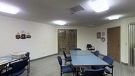 This single-floor space functions as a dedicated meeting or community room, thoughtfully laid out to accommodate group gatherings and collaborative activities. The room's centerpiece is a large rectangular conference table surrounded by several padded blue chairs, offering ample seating for meetings, discussions, or events. Overhead, flush-mounted fluorescent light panels provide bright, even illumination throughout the space, complementing the neutral, light-colored walls and textured ceiling.

A standout architectural feature is the pair of classic wood-framed French doors with grid-pane glass inserts, which serve as the main entry point and lend a warm, traditional character to the otherwise functional interior. The carpeted flooring adds a degree of comfort underfoot while contributing to a quieter acoustic environment.

Along the walls, subtle decorative touches — including wall clocks and small ornamental accents — give the space a welcoming, community-oriented feel. A standalone chair positioned near the wall offers additional flexible seating. HVAC vents integrated into the ceiling ensure a comfortable climate year-round. Overall, the room balances practicality with a modest, inviting ambiance well-suited for regular use as a meeting, training, or multipurpose gathering space.