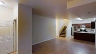 This two-story residence offers a straightforward and functional layout designed to maximize usable living space across both floors. The main level features an open living area finished with warm-toned, hardwood-style flooring that extends throughout, providing a cohesive and easy-to-maintain surface underfoot. Neutral-colored walls throughout the space create a clean, versatile backdrop suitable for a variety of interior styles and décor preferences.

A carpeted staircase, positioned centrally within the main level, connects the two floors and serves as a natural focal point of the lower level. Adjacent to the staircase, a doorway provides access to an additional room or utility space. White baseboards throughout offer a subtle contrast to the flooring and walls, adding a clean, finished look to the interior.

Partially visible at the edge of the main level is what appears to be a kitchen area, suggesting an open or semi-open floor plan on the ground floor. The overall ambiance of the space is calm and understated, with a neutral palette that lends itself well to personalization.