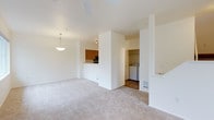 This two-floor residence is designed with an open-concept layout that maximizes the sense of space across the main living area. The living room is finished with light beige carpeting and bright white walls, creating a clean and neutral aesthetic. A ceiling fan overhead provides both air circulation and subtle visual interest, while a pendant light fixture near the kitchen adds warm, focused illumination.

The kitchen is partially open to the living area, separated by a half-wall that also functions as a breakfast bar. Warm wood-toned cabinetry lines the kitchen walls, complemented by a built-in microwave and dark countertops, offering a functional and efficient cooking space.

Adjacent to the kitchen, a dedicated in-unit laundry area is conveniently accessible, housing a full-sized washer and dryer. This practical space is tucked neatly behind a structural partition, keeping it separate from the main living zone while remaining easily reachable.

The overall ambiance of the main level is light and airy, with consistent neutral tones throughout. A fireplace mantle is visible along one wall, adding a subtle architectural detail. The two-floor layout suggests additional living or sleeping spaces above, contributing to the home's versatility and functional depth.