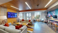 Located at 7004 Tavistock Lakes Blvd in Orlando's Lake Nona neighborhood, this second lobby lounge is a thoughtfully designed communal space that blends comfort with vibrant, contemporary style. The main lounge area features plush neutral sofas accented with bold orange and red throw pillows, arranged around a wood coffee table atop a colorful multi-striped area rug. A large wall-mounted TV, warm wood-paneled ceiling with recessed linear lighting, and expansive windows flooding the space with natural light create an inviting, lively atmosphere. Adjacent to the lounge is a dedicated game and activity area with wooden tables and black wishbone chairs, board games including Monopoly and checkers, and a wall-mounted TV beneath a striking branching chandelier. Large floor-to-ceiling windows offer bright outdoor views throughout. A built-in kitchenette serves the space, featuring dark countertops, a stainless steel dishwasher, and a sink framed by a distinctive illuminated Lake Nona map graphic and vertical wood slat accents. A secluded reading nook provides an intimate retreat, with a built-in upholstered bench set within a wood-framed alcove featuring geometric patterned wallpaper, open shelving, and a bold red accent wall. Polished concrete floors tie the entire space together, reflecting the abundant natural light throughout.