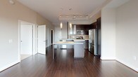 This single-floor residence is designed with a contemporary aesthetic, combining functional layout with refined finishes throughout. The open living area is a standout feature, showcasing rich dark hardwood flooring and a dramatic wall of floor-to-ceiling windows that flood the space with natural light and frame sweeping outdoor views of the surrounding skyline and treetops. A sliding glass door provides direct access to a private balcony, extending the living space outdoors.

The kitchen is well-appointed and thoughtfully designed, featuring dark espresso cabinetry paired with light quartz countertops, a tiled backsplash, and a full suite of stainless steel appliances including a French door refrigerator, built-in microwave, range, and dishwasher. A central island with seating offers additional prep space and casual dining. Pendant lighting and a tray ceiling with track lighting above add a polished finishing touch.

The bedroom continues the bright, airy feel with large sliding glass doors opening to the balcony and plush carpeting underfoot. The bathroom is cleanly finished with white tile flooring, a soaking tub and shower combination accented by a vertical strip of warm brown tile, a dark vanity, and white countertop. Throughout, the unit maintains a cohesive, modern palette.