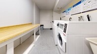 Located at 95 Generation Blvd in Toronto's Scarborough district, this shared laundry facility is a efficiently designed, single-room space that serves its users with a practical and well-organized layout. The room is finished with crisp white walls and a smooth concrete floor, giving it a clean, utilitarian aesthetic throughout.

Along one wall, a row of commercial-grade top-load and front-load washing machines and dryers are neatly arranged, providing ample laundry capacity. Instructional signage is posted above the machines for user guidance. A freestanding utility sink with a faucet is positioned nearby, offering a convenient handwashing or pre-treatment station.

Opposite the appliances, a sturdy built-in folding table with a warm wood-toned butcher block surface runs the length of the room, supported by a white metal frame with integrated lower shelving. This generous surface provides practical space for sorting, folding, and organizing laundry. A metal waste bin is also conveniently placed within the room.

Overhead, fluorescent strip lighting runs along the ceiling, ensuring the space is uniformly and brightly lit. An HVAC vent provides climate control. Access to the room is through a solid door, and an adjacent alcove offers additional utility space. Overall, the facility is clean, functional, and well-equipped for communal use.