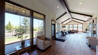 Located at 201 Deann Dr in Independence, Oregon, this single-story space showcases a harmonious blend of rustic charm and modern design. The interior is immediately striking, defined by soaring vaulted ceilings accented with rich, dark-stained exposed wood beams that run the length of the building, creating a dramatic yet welcoming atmosphere throughout.

Rich hardwood flooring flows seamlessly from room to room, grounding the open layout with warmth and continuity. The main living area is anchored by a generous sectional sofa, a wood-framed coffee table, and a stone fireplace that adds a classic, cozy focal point. A large bank of windows along the rear wall floods the space with natural light and frames unobstructed views of the surrounding landscape.

A central corridor opens into multiple seating areas, offering flexible arrangements of armchairs and sofas suited for gathering or quiet relaxation. A dining area with a sturdy table and industrial-style chairs provides additional functionality. The fully equipped kitchen features slate-gray cabinetry, dark granite countertops, stainless steel appliances including a French door refrigerator and dishwasher, and a stone accent wall beside the fireplace. Wood-framed glass doors throughout enhance the connection between indoor and outdoor spaces, completing this inviting and well-appointed environment.