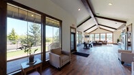 Located at 201 Deann Dr in Independence, Oregon, this single-story space showcases a harmonious blend of rustic charm and modern design. The interior is immediately striking, defined by soaring vaulted ceilings accented with rich, dark-stained exposed wood beams that run the length of the building, creating a dramatic yet welcoming atmosphere throughout.

Rich hardwood flooring flows seamlessly from room to room, grounding the open layout with warmth and continuity. The main living area is anchored by a generous sectional sofa, a wood-framed coffee table, and a stone fireplace that adds a classic, cozy focal point. A large bank of windows along the rear wall floods the space with natural light and frames unobstructed views of the surrounding landscape.

A central corridor opens into multiple seating areas, offering flexible arrangements of armchairs and sofas suited for gathering or quiet relaxation. A dining area with a sturdy table and industrial-style chairs provides additional functionality. The fully equipped kitchen features slate-gray cabinetry, dark granite countertops, stainless steel appliances including a French door refrigerator and dishwasher, and a stone accent wall beside the fireplace. Wood-framed glass doors throughout enhance the connection between indoor and outdoor spaces, completing this inviting and well-appointed environment.