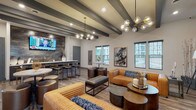 This single-story clubhouse is thoughtfully designed to serve as a premier social and recreational hub. The main lounge area is anchored by warm wood-look flooring, exposed dark beam ceilings, and contemporary globe chandeliers that cast an inviting glow. Richly upholstered caramel leather sofas, tufted benches, and round accent tables create flexible seating arrangements ideal for both casual relaxation and group gatherings. A wall-mounted flat-screen TV backed by a textured stone accent wall adds a focal point to the lounge, while multiple windows allow natural light to fill the space and provide views of the surrounding exterior.

Adjacent to the lounge, a well-appointed wet bar area features dark cabinetry, crisp white countertops, a stainless steel sink, and a bold tile backsplash beneath a second mounted television—perfect for entertaining. A hallway connects to additional rooms and restroom facilities, with wall-mounted TVs placed conveniently throughout.

The game room stands out with soaring ceilings, abundant window coverage, and a full-size billiards table illuminated by a geometric chandelier, creating a bright and energetic atmosphere. Throughout the clubhouse, the consistent use of neutral tones, modern fixtures, and curated artwork contributes to a cohesive, upscale aesthetic.