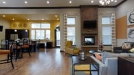 This single-floor amenity space combines comfort and functionality in a thoughtfully designed open layout. The main lounge area is anchored by a striking full-height brick fireplace, complete with a wood mantle and wall-mounted television above, serving as a natural focal point for the room. Rich hardwood flooring flows throughout, complementing the warm, neutral tones of the walls accented by bold yellow paint in the adjoining area. Seating arrangements include a yellow accent armchair and a plush sofa, creating an inviting atmosphere for relaxation and socializing. Large windows fitted with classic white blinds and patterned curtain panels flood the space with natural light, enhancing the open, airy feel. Recessed lighting adds to the bright, well-illuminated environment. Adjacent to the lounge, a business or co-working area features bar-height countertops, high stools, and additional windows with exterior views, making it an ideal setting for productivity. A pendant light fixture overhead adds a polished finishing touch to this zone. The overall ambiance is warm and contemporary, blending residential comfort with practical community-focused design throughout the space.