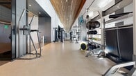 Located at 1177 Market Street in San Francisco's Civic Center neighborhood, this fitness center offers a well-appointed, single-floor facility designed with both function and aesthetics in mind. The space is divided into distinct zones, each purpose-built to support a comprehensive workout experience.

The cardio area is particularly striking, featuring a long, mirror-lined corridor with rows of Assault Fitness rowers, ellipticals, and treadmills. A slatted wood ceiling with integrated linear lighting runs the length of the room, creating a warm contrast against the cool tones of the surrounding walls and polished flooring, lending the space a sense of depth and sophistication.

Adjacent to the cardio zone, the strength training area is well-equipped with cable machines, free weights, medicine balls, TRX suspension systems, and a variety of resistance equipment organized along mirrored walls. A glass-partitioned studio space provides a bright, open environment for functional training, featuring high ceilings and large windows that allow natural light to fill the room with views of the surrounding urban streetscape.

The overall design blends natural wood accents with dark architectural glass partitions, light-toned cork and hardwood flooring in a herringbone pattern, and bold red accent seating — creating a polished, modern fitness environment suited for a wide range of training disciplines.