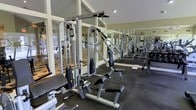 The fitness center at Spring House On The Park is a well-appointed, single-level space designed to support a comprehensive workout routine. The room is outfitted with a variety of strength training machines, including cable pulley systems, adjustable benches, and weight stack equipment, all arranged to allow comfortable movement between stations. Rubber flooring provides a durable, slip-resistant surface throughout the workout area. Large windows along the perimeter invite abundant natural light, creating an open and energizing atmosphere. Full-length mirrored walls visually expand the space while allowing users to monitor their form during exercise. The vaulted ceiling adds a sense of height and airiness, preventing the room from feeling confined despite its focused purpose. Framed by white-trimmed windows and neutral walls, the overall aesthetic is clean and functional. The layout reflects thoughtful planning, offering enough space between equipment for safe and efficient use. This fitness center serves as a convenient, well-maintained amenity for residents of Spring House On The Park.