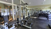 The fitness center at Spring House On The Park is a well-appointed, single-level space designed to support a comprehensive workout routine. The room is outfitted with a variety of strength training machines, including cable pulley systems, adjustable benches, and weight stack equipment, all arranged to allow comfortable movement between stations. Rubber flooring provides a durable, slip-resistant surface throughout the workout area. Large windows along the perimeter invite abundant natural light, creating an open and energizing atmosphere. Full-length mirrored walls visually expand the space while allowing users to monitor their form during exercise. The vaulted ceiling adds a sense of height and airiness, preventing the room from feeling confined despite its focused purpose. Framed by white-trimmed windows and neutral walls, the overall aesthetic is clean and functional. The layout reflects thoughtful planning, offering enough space between equipment for safe and efficient use. This fitness center serves as a convenient, well-maintained amenity for residents of Spring House On The Park.