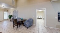 Located at 1919 NW 142nd St in Oklahoma City, Oklahoma, this single-floor space offers a thoughtfully arranged layout with two living areas, a full kitchen, and a warm, inviting ambiance throughout. The main living area immediately impresses with dramatically vaulted ceilings that amplify the sense of openness, complemented by light neutral walls and consistent light tile flooring. A reception-style counter separates the entry zone from the main seating area, where large windows allow natural light to filter in. A second living area, accessible through elegant French doors, provides a quieter retreat with carpeted flooring and dual windows fitted with blinds. The well-equipped kitchen features rich medium-toned wood cabinetry, granite-style countertops, stainless steel appliances, and crown molding detailing, conveying both function and style. A cozy dining nook with a round table and eclectic seating sits adjacent to the kitchen, opening toward an additional utility room. Decorative touches throughout — including large-scale artwork, a rustic distressed cabinet piece near the entry, and mirrored accent furniture — lend the space a curated, welcoming character. A dedicated workspace with a desk and artwork rounds out the layout, making this a versatile, multi-functional space suited for a variety of uses.