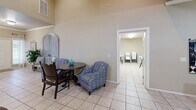 Located at 1919 NW 142nd St in Oklahoma City, Oklahoma, this single-floor space offers a thoughtfully arranged layout with two living areas, a full kitchen, and a warm, inviting ambiance throughout. The main living area immediately impresses with dramatically vaulted ceilings that amplify the sense of openness, complemented by light neutral walls and consistent light tile flooring. A reception-style counter separates the entry zone from the main seating area, where large windows allow natural light to filter in. A second living area, accessible through elegant French doors, provides a quieter retreat with carpeted flooring and dual windows fitted with blinds. The well-equipped kitchen features rich medium-toned wood cabinetry, granite-style countertops, stainless steel appliances, and crown molding detailing, conveying both function and style. A cozy dining nook with a round table and eclectic seating sits adjacent to the kitchen, opening toward an additional utility room. Decorative touches throughout — including large-scale artwork, a rustic distressed cabinet piece near the entry, and mirrored accent furniture — lend the space a curated, welcoming character. A dedicated workspace with a desk and artwork rounds out the layout, making this a versatile, multi-functional space suited for a variety of uses.